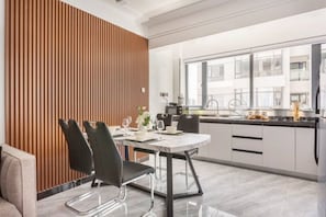 A modern kitchen with a dining area, featuring a wooden panel wall, white cabinets, and a large window. - Inikas Living_Luxurious 1 bedroom apartment  (Nairobi)