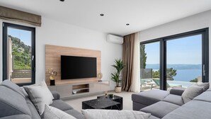 A modern living room with a large flat-screen TV mounted on a wooden panel, grey sofas, a marble coffee table, and a view of the sea through large windows.