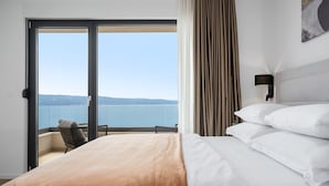A hotel room with a bed, a view of the sea, a balcony with a chair, and a wall-mounted lamp.