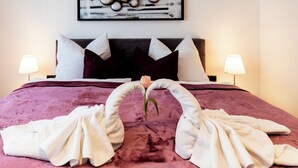 Un lit recouvert d’une couvre-lit violette, deux serviettes blanches disposées en forme de cœur et une seule rose.