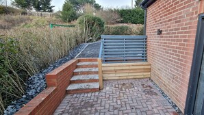 Une terrasse en briques avec un escalier en bois menant à une zone aménagée, le tout entouré de végétation et d’un mur en briques.