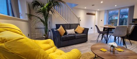 Una sala de estar moderna con un sofá gris oscuro, un sillón amarillo y una mesa de centro de madera. Se ve una escalera con barandillas blancas al fondo.