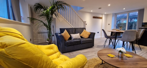 A modern living room with a dark grey sofa, yellow armchair, and a wooden coffee table. A staircase with white railings is visible in the background.