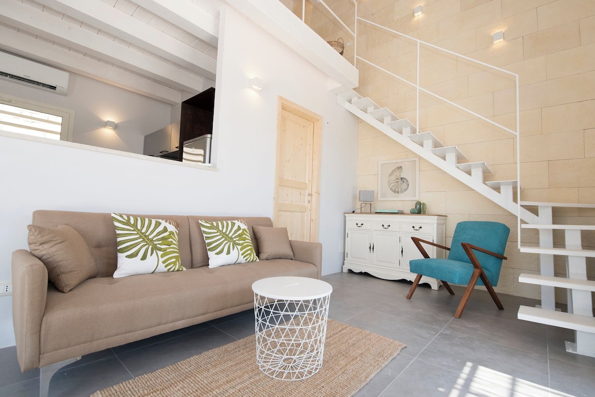 A modern living room with a sofa, a round coffee table, a blue armchair, and a staircase with a white railing.