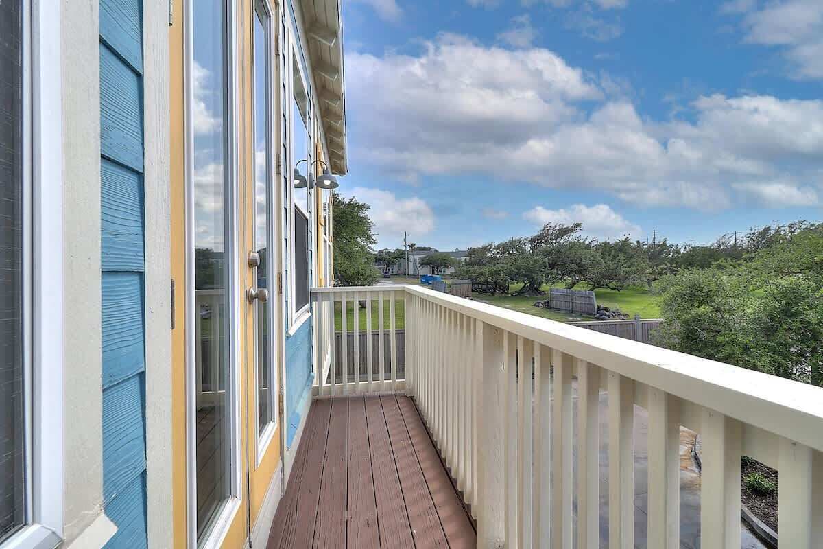 Ein Balkon mit Holzdeck, weißer Geländer und Blick auf eine grüne Landschaft mit Bäumen und blauem Himmel.