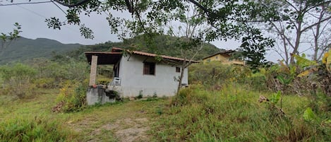 A small, single-story building with a sloped roof, surrounded by dense vegetation and hills in the background.