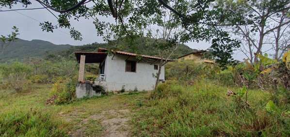 A small, single-story building with a sloped roof, surrounded by dense vegetation and hills in the background.
