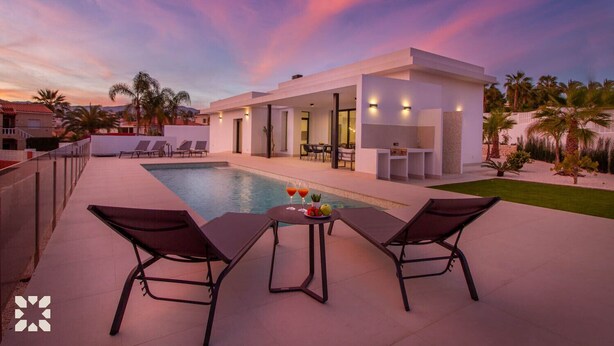 Un área de piscina moderna al aire libre con sillones de descanso y una mesa con bebidas, rodeada por un jardín cuidado y una vista despejada del atardecer.