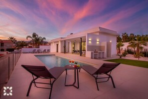 Un área de piscina moderna al aire libre con sillones de descanso y una mesa con bebidas, rodeada por un jardín cuidado y una vista despejada del atardecer.