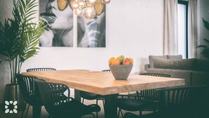 Un comedor con una mesa de madera, sillas negras, un cuenco de fruta, una lámpara colgante moderna y un cuadro en la pared.