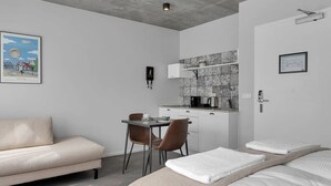 Quarto de hotel moderno com sofá, mesa de jantar com cadeiras, cozinha compacta e cama com toalhas.