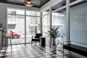 A modern interior with a checkered floor, a bar area with stools, a seating area with a chair, and a large window offering a view of the street.