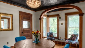 Un comedor con una mesa redonda de madera, sillas azules y un jarrón con flores. Se observan grandes ventanales con vistas a árboles y una lámpara de araña.