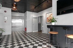 Welcome to our lobby, featuring a checkered floor, a reception desk with seating, a mounted TV, a potted plant, and an elevator.