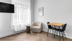 A room with a wooden herringbone floor, a round wooden side table, a beige armchair, a wooden table with two black chairs, a large window with sheer curtains, and a framed picture on the wall. - Swan Stylish Double Studio (Reykjavík)
