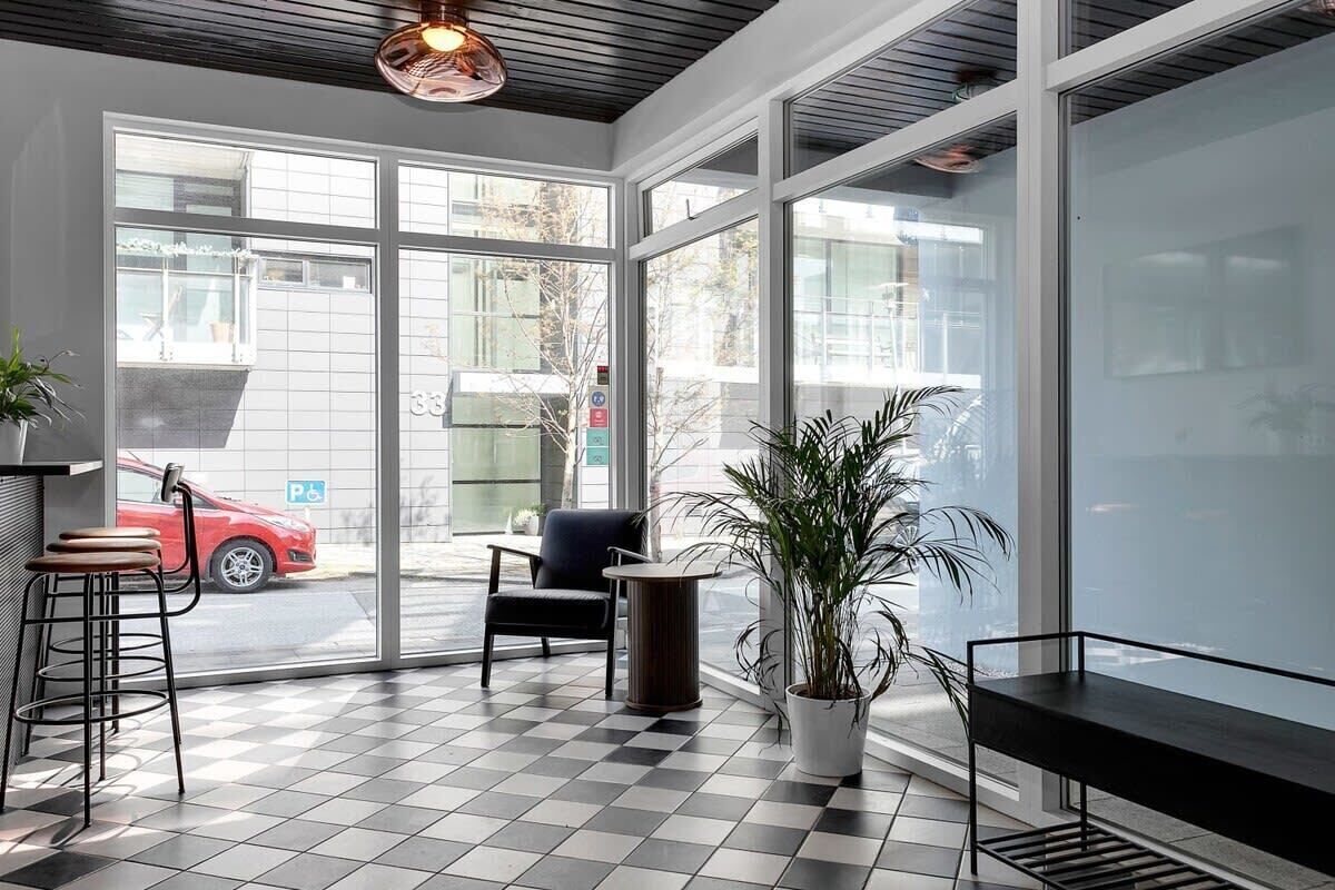 A modern interior with a checkered floor, a bar area with stools, a seating area with a chair, and a large window offering a view of the street.