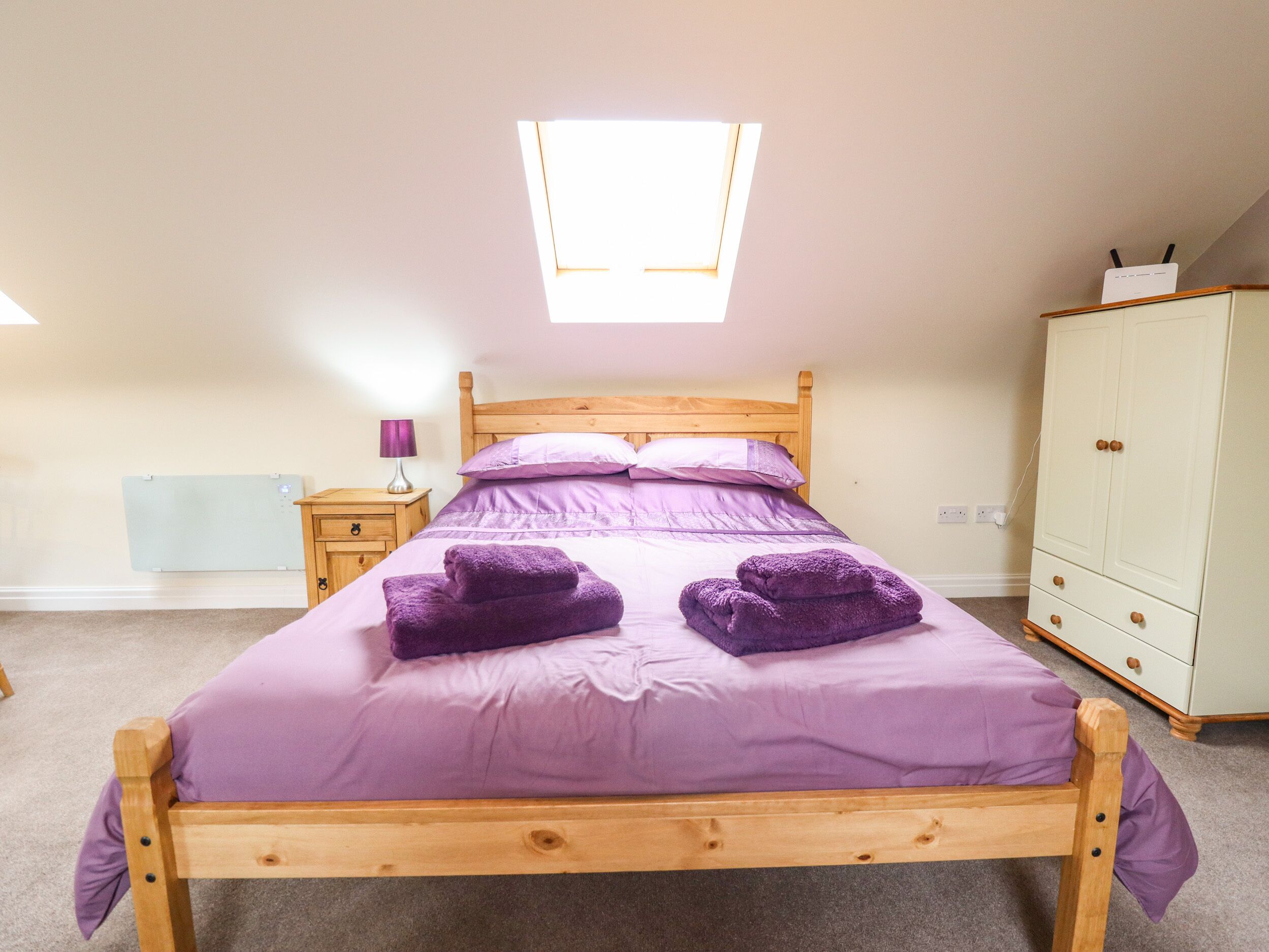 A bedroom with a wooden bed, purple bedding, a nightstand, a lamp, a wardrobe, and a skylight.
