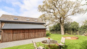 Une grange en bois avec un toit en métal, un chemin de gravier et une zone herbeuse où se trouvent une table ronde et deux chaises.
