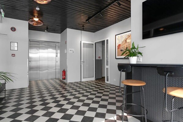 Un hall d’entrée avec un sol à carreaux, un comptoir de réception avec des tabourets, une plante en pot et un extincteur.