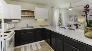 A modern kitchen with black cabinets, a white countertop, a microwave, and a stove. There is a dining area with a table and chairs, and a ceiling fan. - Resort Living on the Coconut Coast (Kapaa)