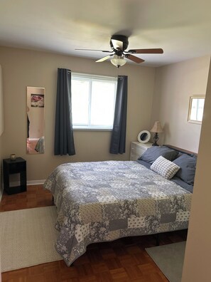 Un dormitorio con cama, ventilador de techo, ventana con cortinas, lámpara de noche, mesita de noche y espejo.