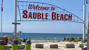 Une plage avec un panneau indiquant « Bienvenue à Sauble Beach », un ciel dégagé et l’océan en arrière-plan.