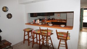 A compact kitchen with a central island, four wooden bar stools with colorful cushions, and a refrigerator. - Pelican Inlet C218 2 Bedroom Condo by RedAwning (St. Augustine)