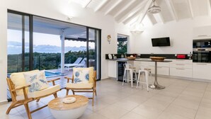 A modern kitchen with a large island, a dining area with white chairs, and a view of a pool and mountains through large sliding glass doors. - Superbe Villa vue mer (Sainte Anne)