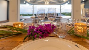 A dining table set with white plates, clear glasses, and candles, with a view of the outdoors. - Superbe Villa vue mer (Sainte Anne)