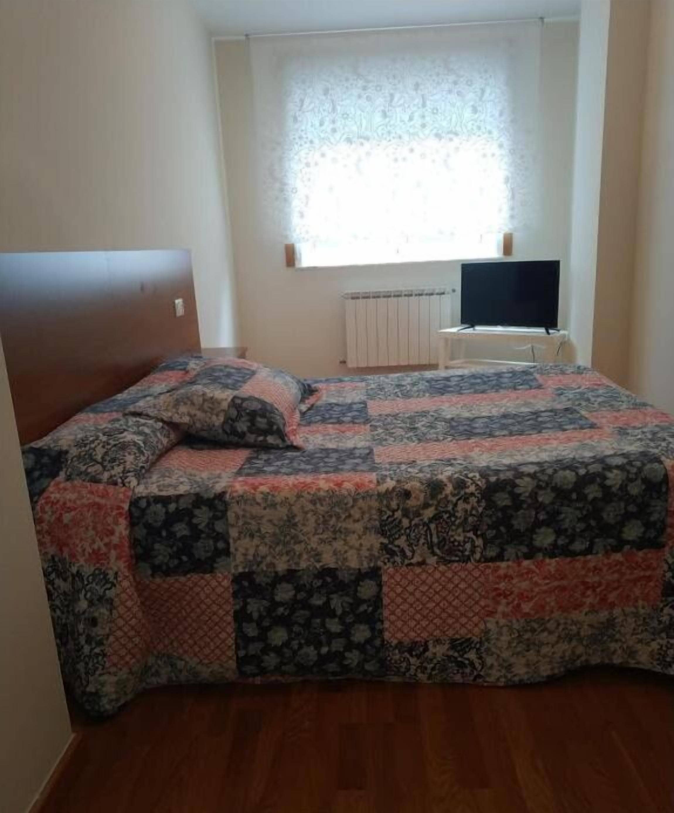 A bedroom with a wooden headboard, a quilted bedspread, a television, and a window with a patterned curtain.