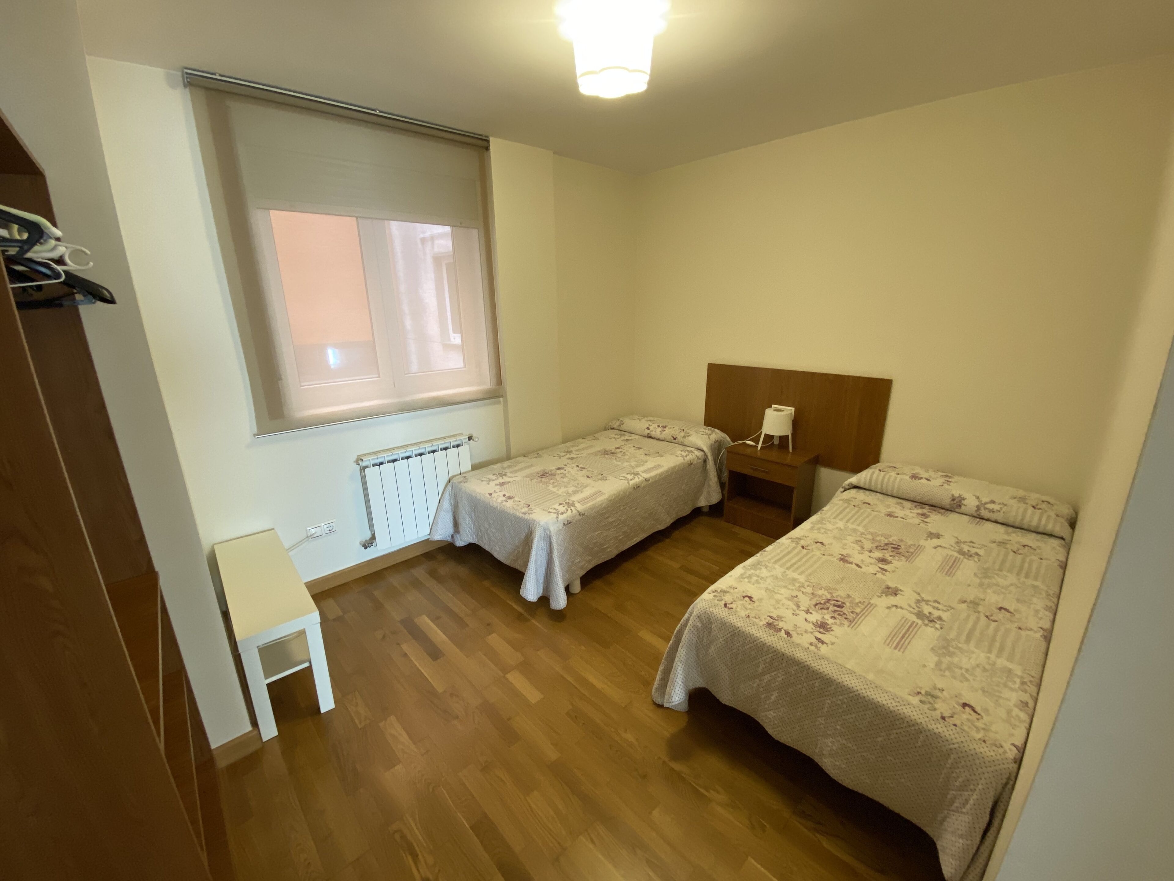 Ein Zimmer mit zwei Einzelbetten, Holzboden, Heizkörper, einem kleinen Tisch und einem Fenster mit Jalousien.
