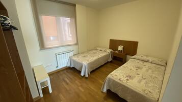 Ein Zimmer mit zwei Einzelbetten, Holzboden, Heizkörper, einem kleinen Tisch und einem Fenster mit Jalousien.
