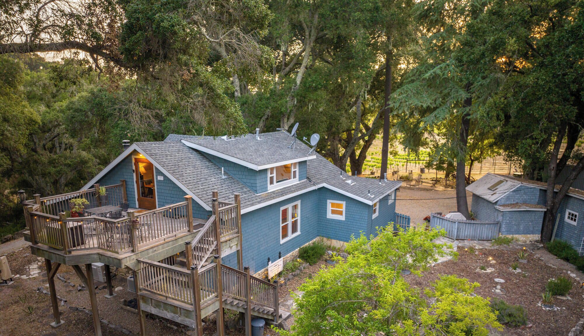 A two-story cabin with a deck, surrounded by trees and a vineyard.