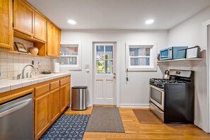A compact kitchen with wooden cabinets, a stainless steel dishwasher, a sink with a faucet, a trash can, a stove, and a microwave.