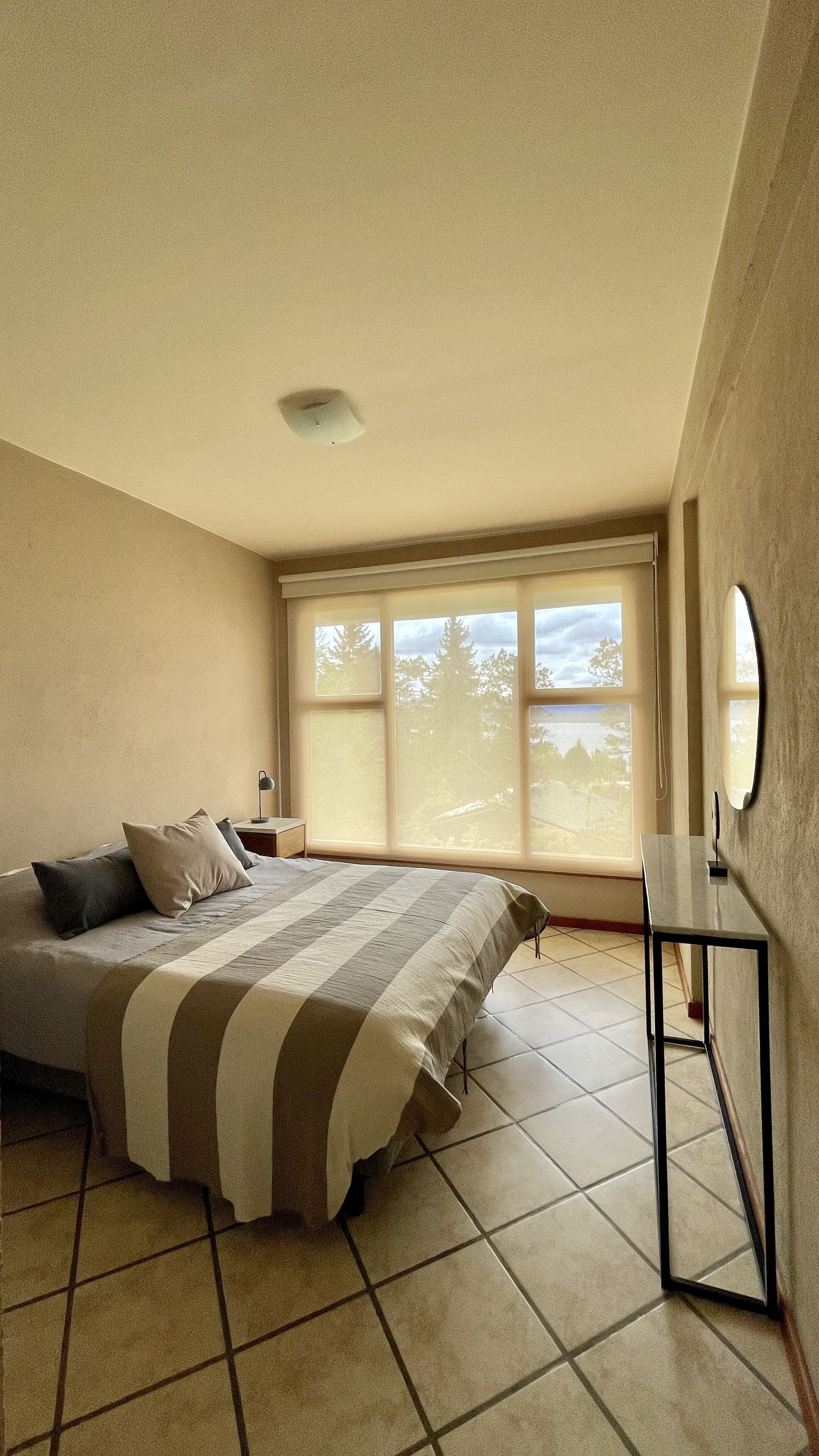 A bedroom with a large bed, a bedside table, a mirror, and a window with a view of trees.