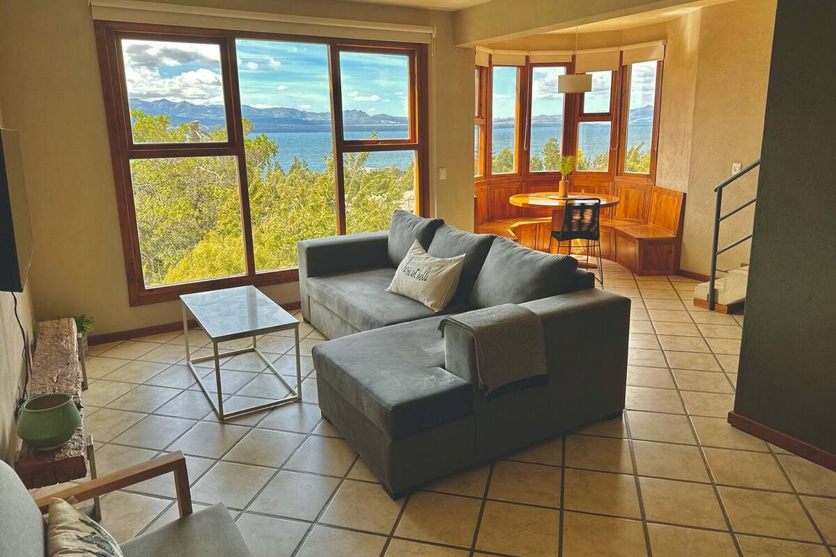 Amplia sala de estar con un sofá gris, una mesita de centro y grandes ventanas con vistas a montañas y árboles.