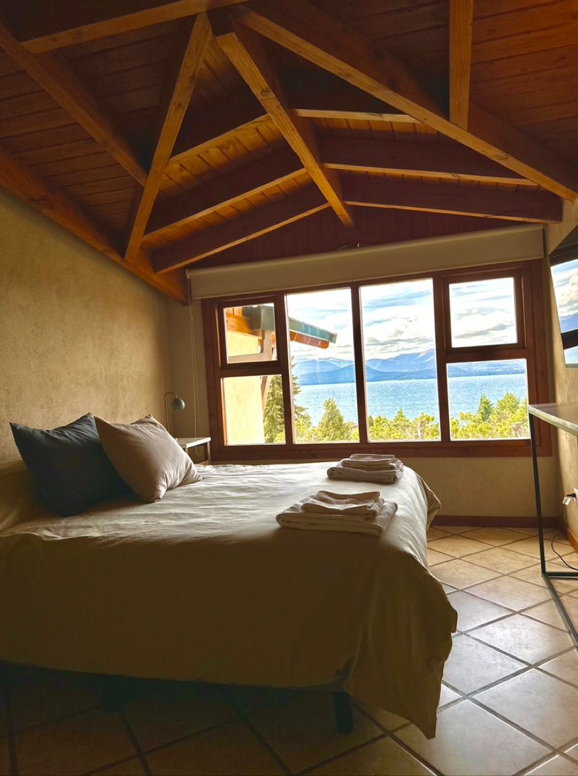 A bedroom with a bed, pillows, a window with a view of mountains and water, and a wooden ceiling.