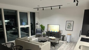 Una sala moderna con sofá, mesa de centro y televisor. Hay un comedor con sillas y una mesa. La habitación tiene ventanas amplias y vista a la ciudad de noche.