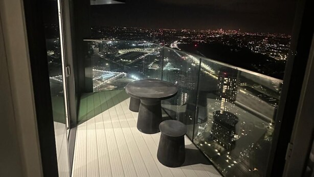 Un balcón con mesa y dos taburetes, con vistas a un paisaje urbano nocturno.