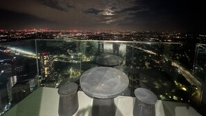 Habitación en un rascacielos con vista panorámica de la ciudad de noche, que cuenta con barandilla de vidrio y una mesa redonda con taburetes.
