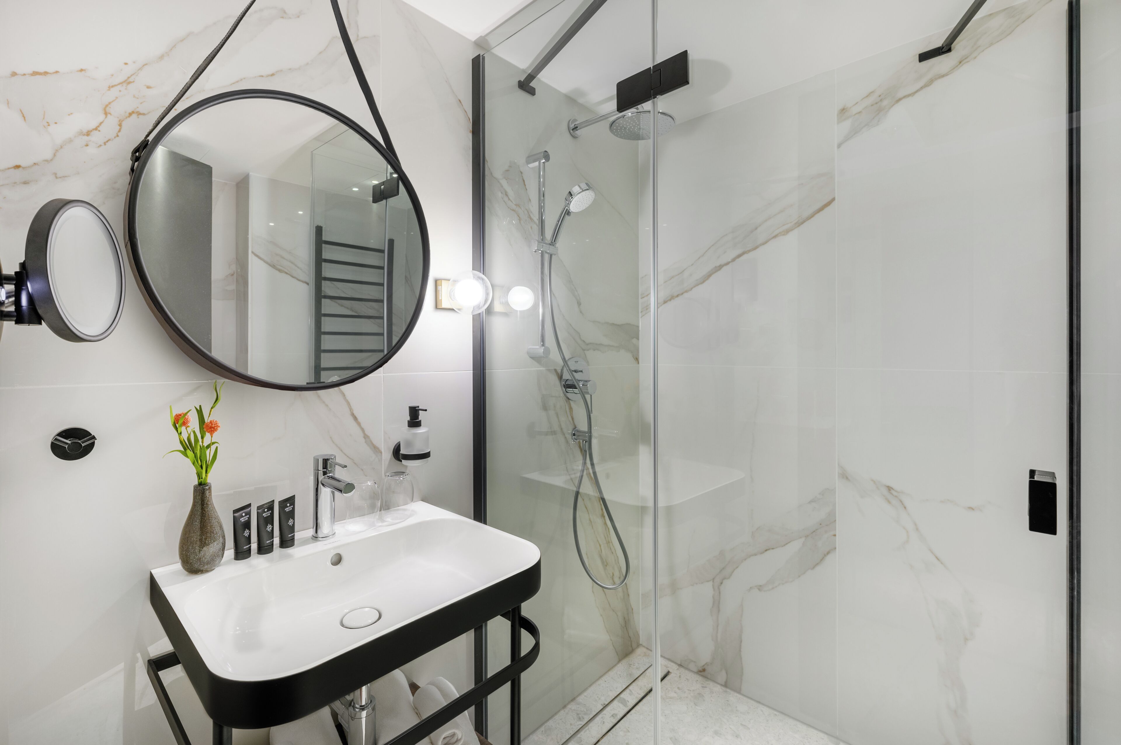 A modern bathroom with a glass-enclosed shower, a round mirror, a white sink, and a vase with flowers.