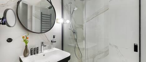 A modern bathroom with a glass-enclosed shower, a round mirror, a white sink, and a vase with flowers.