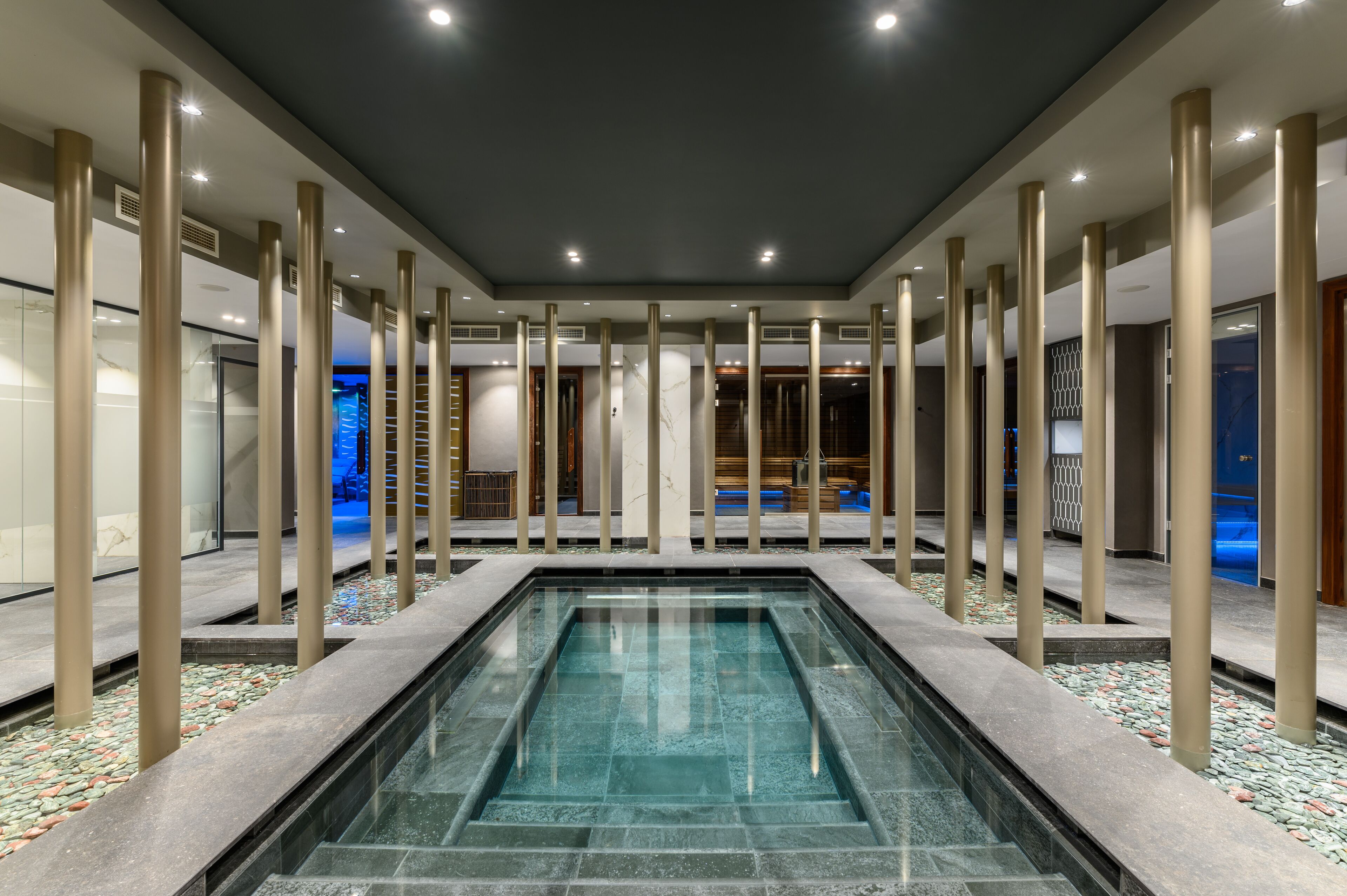 Un área de piscina interior moderna con una piscina rectangular, pilares color beige y suelo de baldosas.