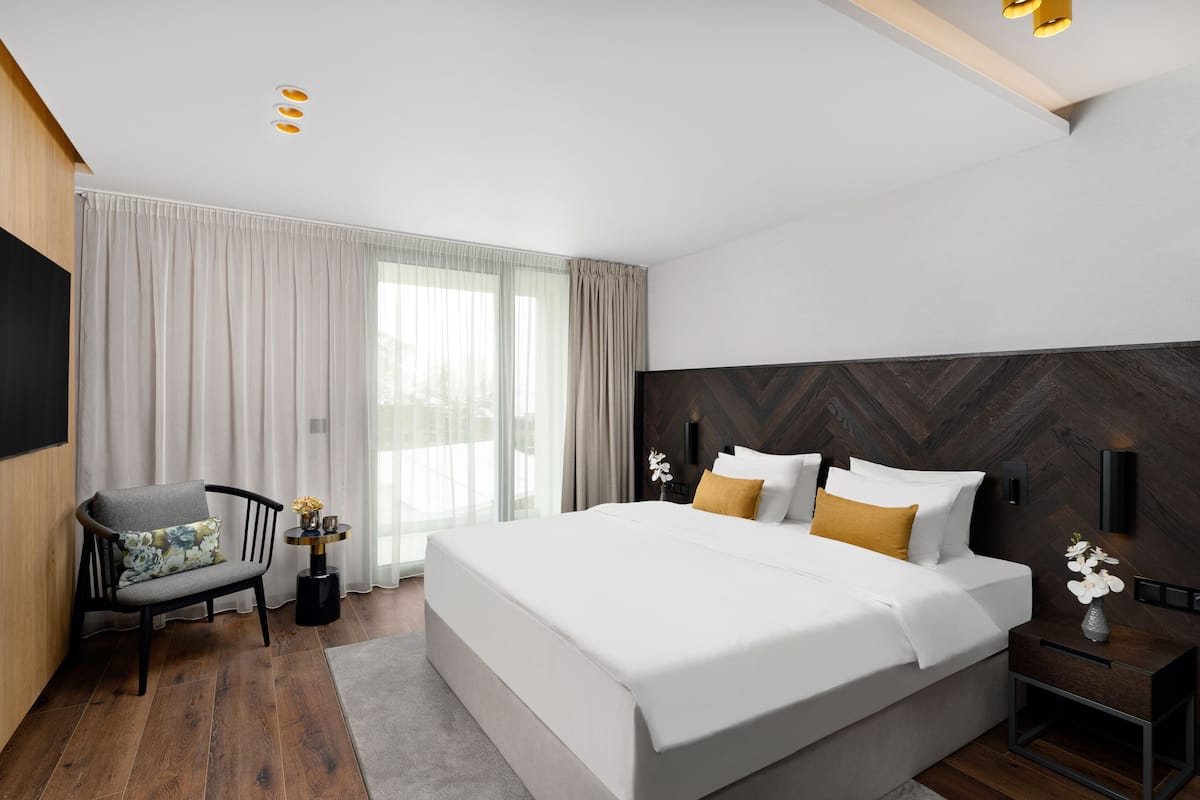 A modern hotel room with a large bed, a seating area with a chair, a small table with a vase, and a large window with curtains.