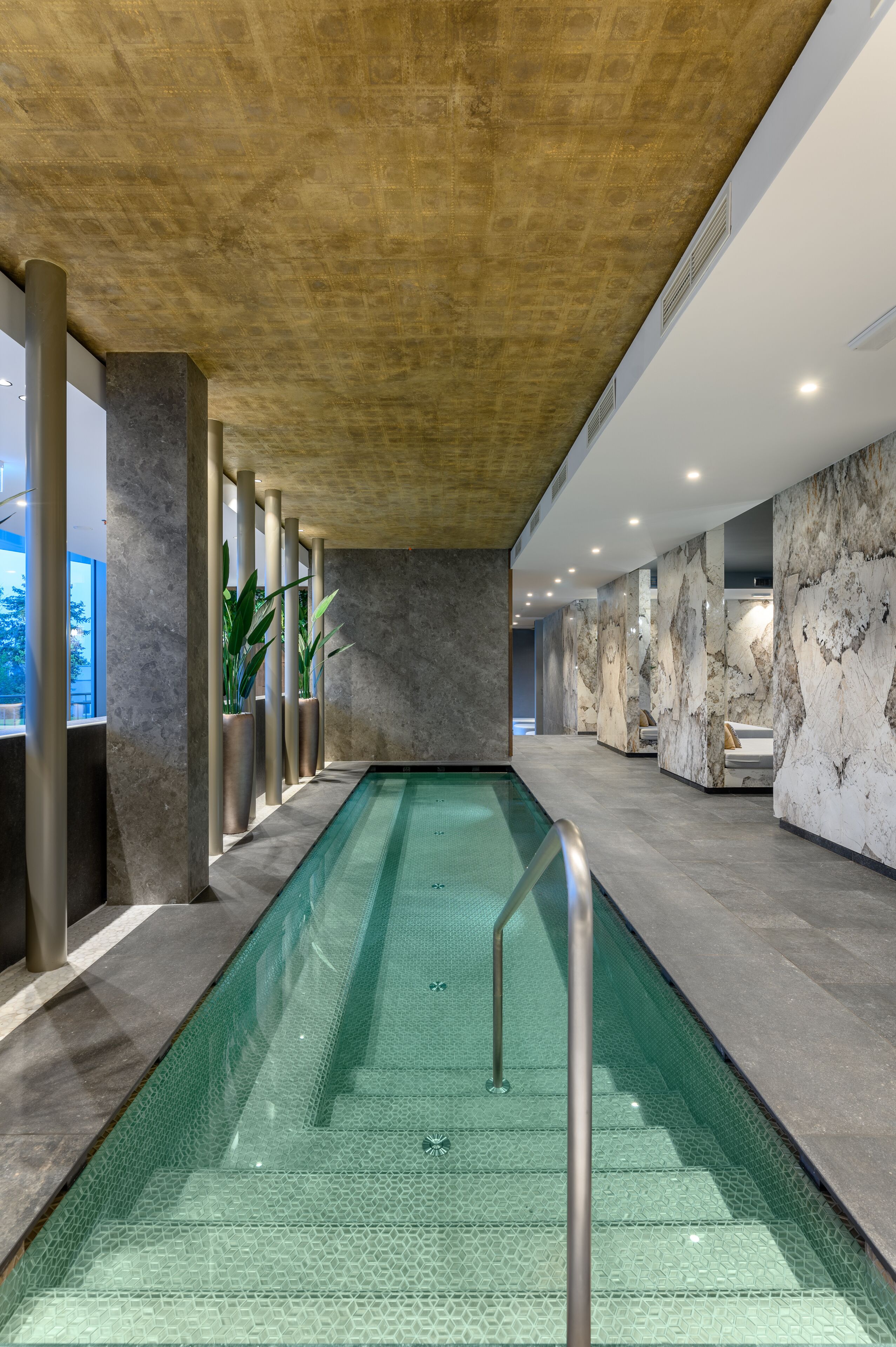 A modern indoor swimming pool with a glass bottom, surrounded by marble walls and columns, and a wooden ceiling.