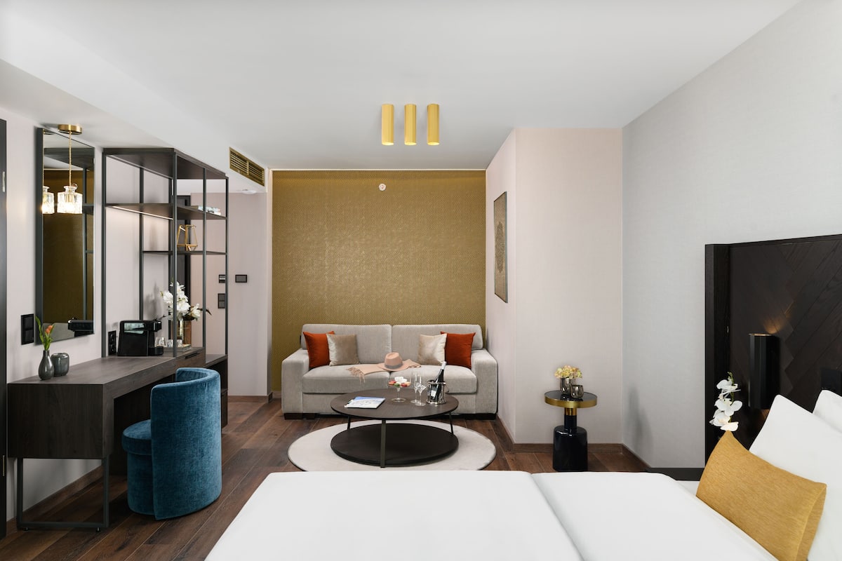 A modern hotel room with a sofa, a round coffee table, a desk with a chair, and a bed.