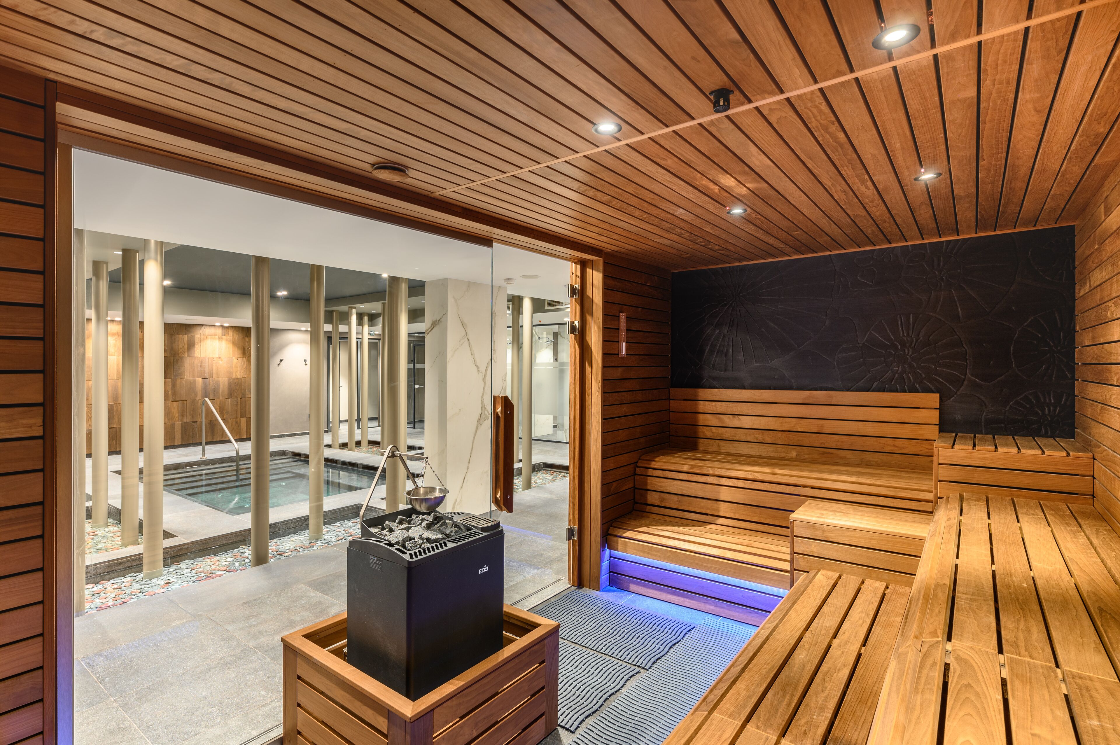 Un sauna avec des bancs en bois et un mur en pierre, une porte en verre donnant sur un espace piscine, et un radiateur noir au centre.