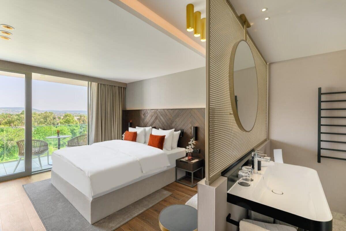 A modern hotel room with a large bed, a bathroom with a large mirror, and a balcony with a view.