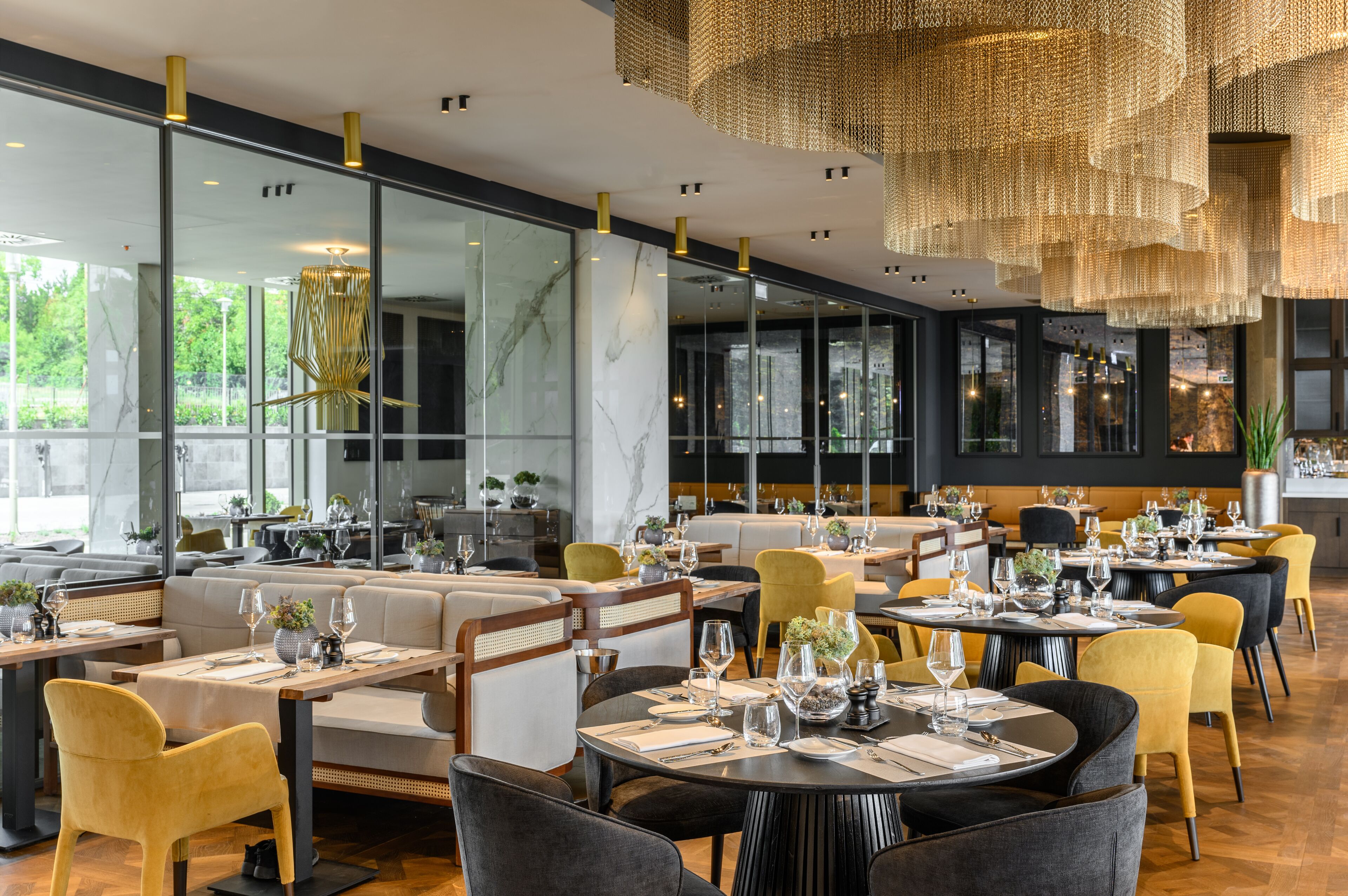 A modern restaurant interior with glass partitions, wooden furniture, and a central chandelier.