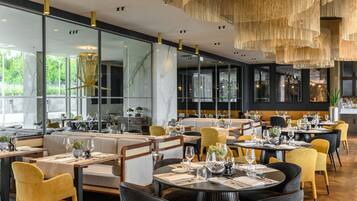 A modern restaurant interior with glass partitions, wooden furniture, and a central chandelier.